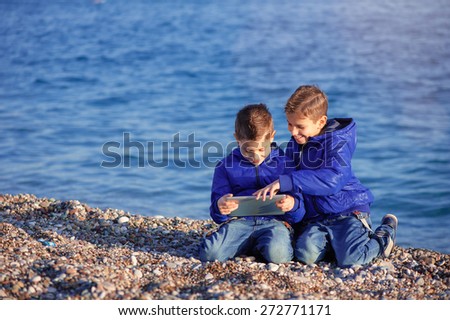 Two happy caucasian kids, brothers, playing together with tablet pc sitting outdoors at pebble beach against the sea