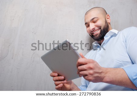 Working on tablet computer. Handsome young arabic man working on digital tablet while leaning on grey wall.