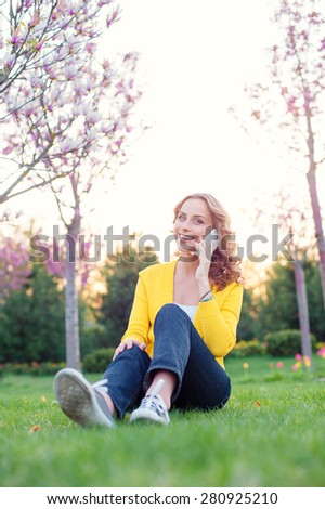 Cheerful conversation. Attractive young woman talking by phone while sitting on grass in spring park.