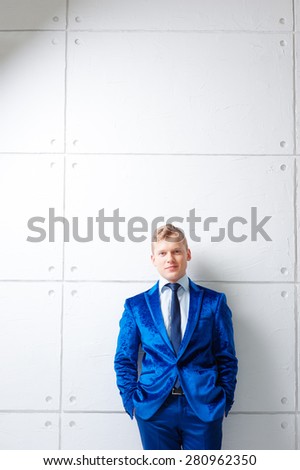 Confident and charisma. Portrait of happy young man in blue suit looking at camera holding hands in pockets while leaning on white wall.