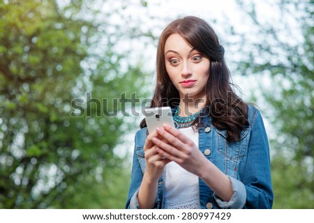 Shocked news. Youth and technology. Amazed young woman using smartphone in green park.
