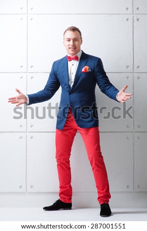 You are welcome! Cheerful young man in jacket and bowtie gesturing welcome sign and smiling while standing against white wall.