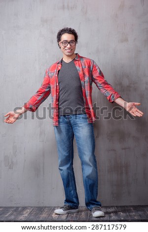 You are welcome! Cheerful Arabic hipster man gesturing welcome sign and smiling while standing against grey background