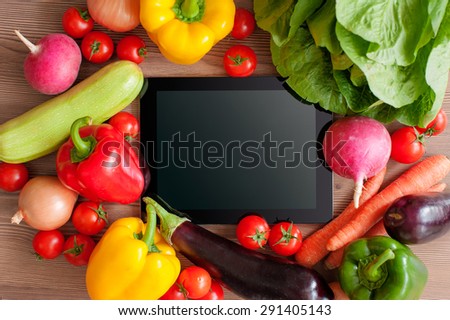 Fresh farm vegetables. Tablet, black screen in the center. Pepper, salad, tomatoes, onion, carrot, eggplant and radish. Concept of digital recipe book. Top view.
