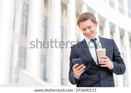 Work and coffee break. Cheerful young businessman using smartphone and holding cup of beverage.