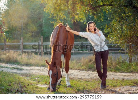 Technology at countryside. Attractive young woman talking by phone while walking with horse outdoors.