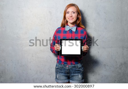 Your text here. Attractive young woman in plaid shirt and bowtie showing screen of tablet computer against grey wall.