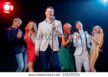 Stand-up comedy and party. Showman with microphone. Group of cheerful friends toasting with wineglasses among confetti.