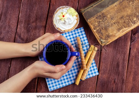Warming hands on a cup of tea. Her hands around the blue cup of tea. A cup of coffee or tea on a wooden table. Antique book, cakes, pastries and a cup of tea.