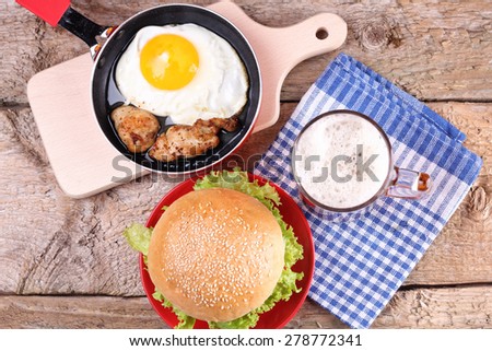 Fried egg and meat on the pan. Roll with sesame and vegetables. Wooden table.  The Burger on the plate. Sandwich. Meat and beer. White fresh bread and vegetables. Alcohol and meat.