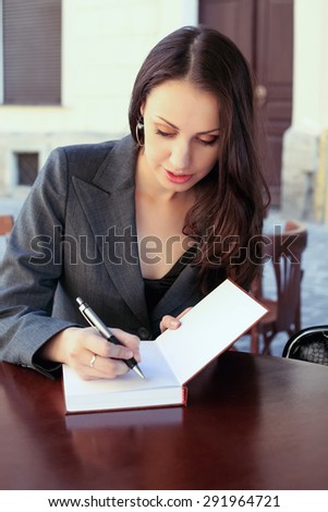 Beautiful confident woman. Woman on the street at the table . Business woman outdoors. Business consultant, to note down, to make notes. Business meeting. Woman with notebook. Business look.