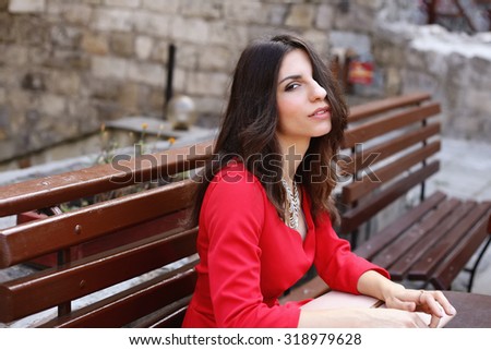 Beautiful girl in the red dress. The girl. sitting on a bench. Outdoor cafe, summer patio. Sexy girl in a scarlet gown. Girl waiting. Girl with necklace. Romantic mood. Love. Resting girl.