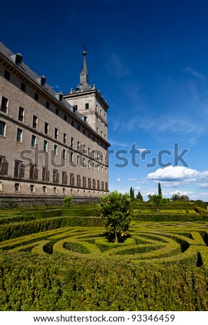 San Lorenzo de El Escorial Monastery with its formal gardens. The towers of the monastery are set of by a bright blue sky with a few white clouds.