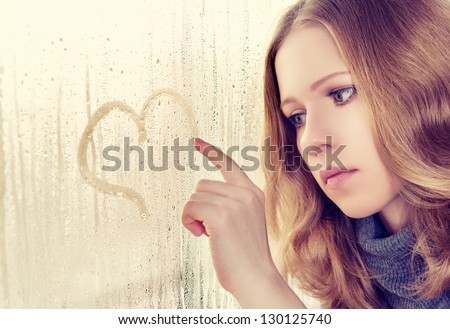 sad enamored girl draws a heart on the window in the rain