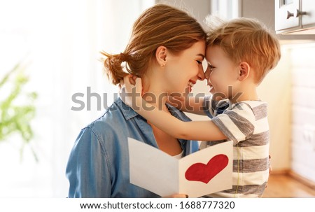 Happy little boy congratulating smiling mother and giving card with red heart during holiday celebration at home