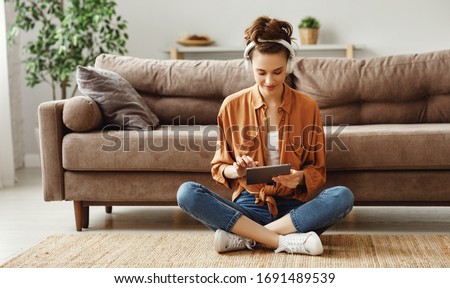Young satisfied female in casual clothes and wireless headphones surfing digital tablet while sitting on floor with legs crossed near soft couch in light modern living room Foto stock © 