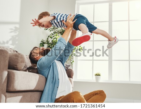 Joyful young man in casual wear sitting on floor and throws up happy little son while having fun together in cozy living room at home