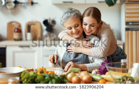 Happy family cheerful young woman embracing mature mother while preparing healthy dish with fresh vegetables in home kitchen