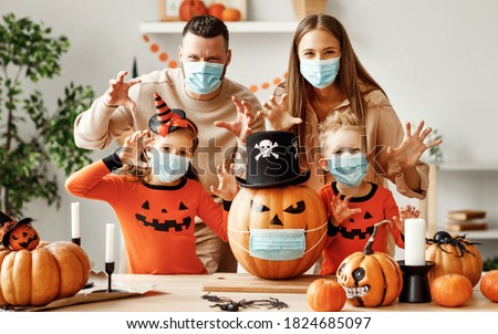 Cheerful family  in medical masks  makes jack o lantern   out of a pumpkin and  scare for camera  in cozy kitchen during Halloween celebration at home during the covid19 coronavirus pandemic