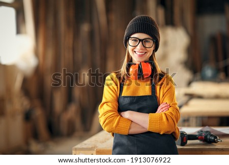 Smiling confident young female joiner in apron standing near workbench and looking at camera friendly while working in craft workshop 商業照片 © 