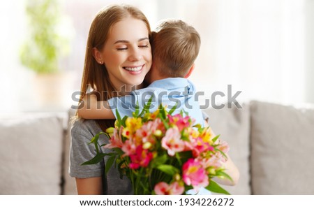Loving little son hugging happy mom and greeting with bouquet of Alstroemerias during Mothers day celebration at home