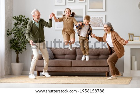 Full length of two excited kids siblings having fun playing with active and positive grandparents in the living room at home, enjoying weekend time with energetic senior old grandma and grandpa