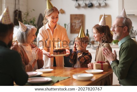 Happy big family gathered in living room table to celebrate birthday of cute little boy, excited child covering eyes with hands while mother surprising her by bringing chocolate cake with lit candles 商業照片 © 