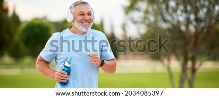 Happy mature man in headphones holding water bottle while jogging outside in park in early morning, senior sportsman running with beaming smile on his face enjoying active healthy lifestyle Foto stock © 