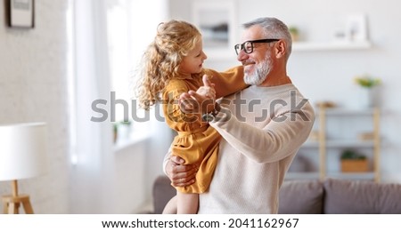 Elegant loving caring grandfather looking at his cute little granddaughter, adorable child girl and positive grandpa holding hands while dancing together in living room at home. Family concept