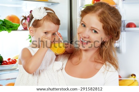 happy family mother and baby daughter drinking orange juice in the kitchen near the refrigerator