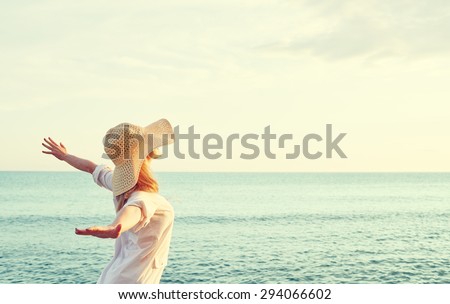 Happy beauty woman in hat is back opened his hands, relaxes and enjoys the sunset over the sea on the beach