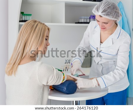 Blood test from vein with beautiful young blond woman by female doctor in white coat medical uniform on the table in white bright room.