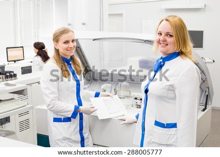 Medical workers women in white uniforms and gloves standing near centrifuge separation machine show information on the paper sheet and smiling.
