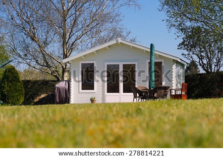 Beautiful luxury wooden Summerhouse in a country garden with tables, chairs, sun umbrella, trees, blue sky, green grass. Summer house is a romantic & relaxing garden retreat.