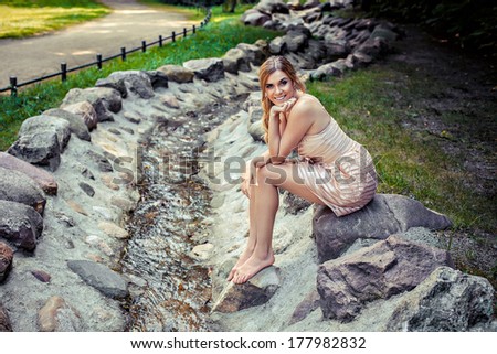 Young beautiful smiling woman sitting on rock by stream in park