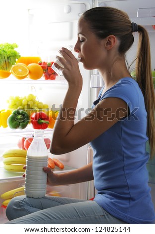Young woman drinking milk near refrigerator at night