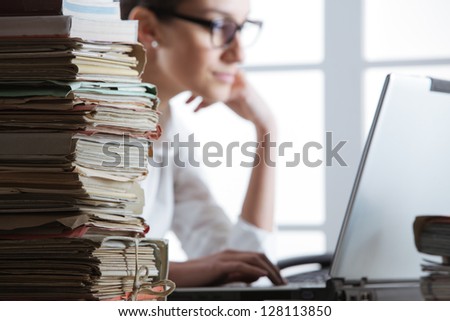 Stack of documents, with secretary who works at the computer in the background