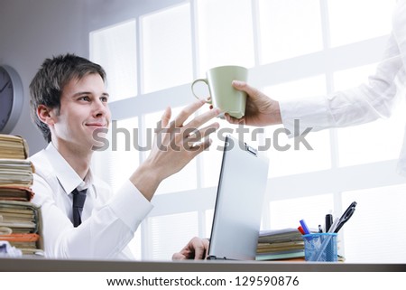 Hand of colleague offering coffee to young man sitting on workplace behind computer