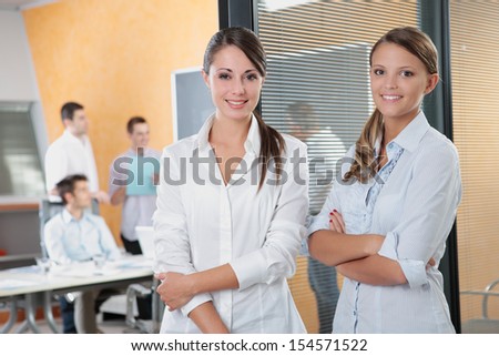 Portrait of two young business woman standing while his team talks in the background