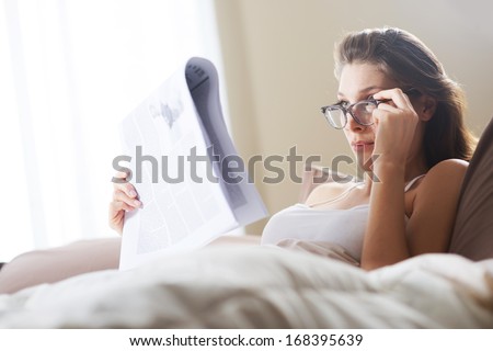 Young woman lying on the bed at home and reading the newspaper