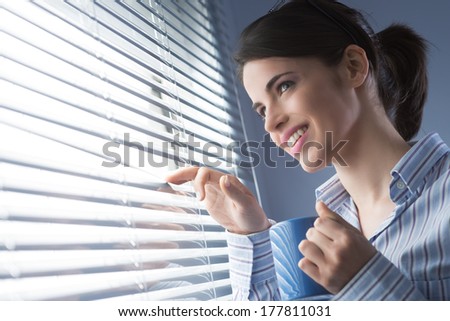Attractive office worker peeking out of blinds and holding a mug.