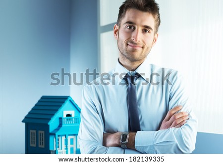 Young real estate agent with arms crossed and model house on background.