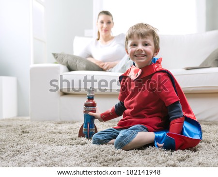 Cute superhero boy paying with toy rocket in the living room with his mother on background.