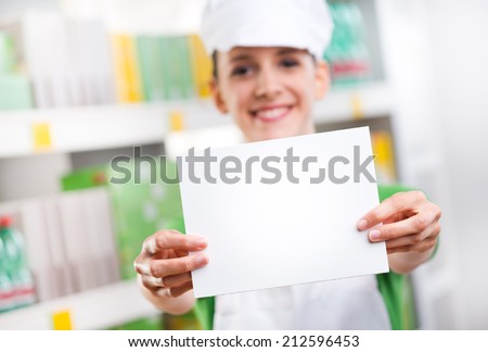 Female sales clerk holding a white card and smiling with supermarket shelf on background.