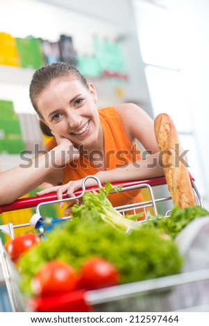 Smiling woman at supermarket with full shopping cart and shelves on background.