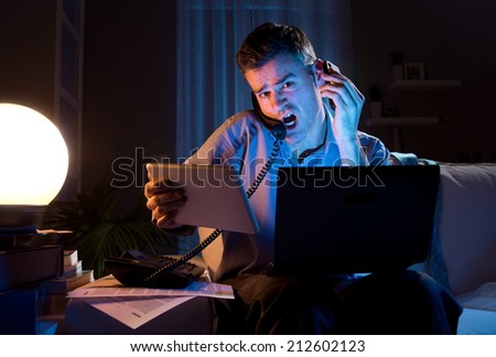 Stressed businessman working overtime late at night in the living room with telephone, computer and tablet.