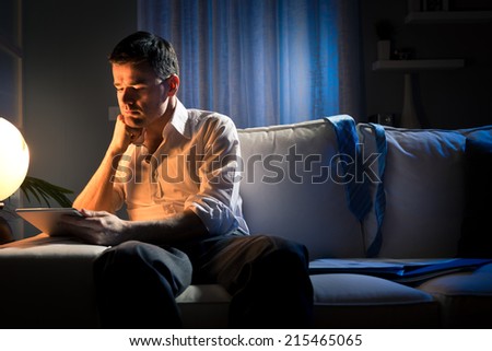 Businessman working overtime late at night in the living room, with tablet and paperwork.