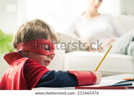 Cute superhero boy drawing with mother reading on background.