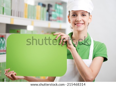 Female sales clerk holding a green sign and smiling with supermarket shelf on background.