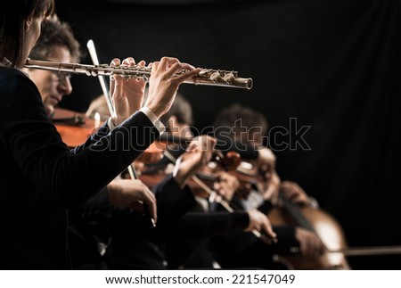 Female flutist close-up with orchestra performing on background.